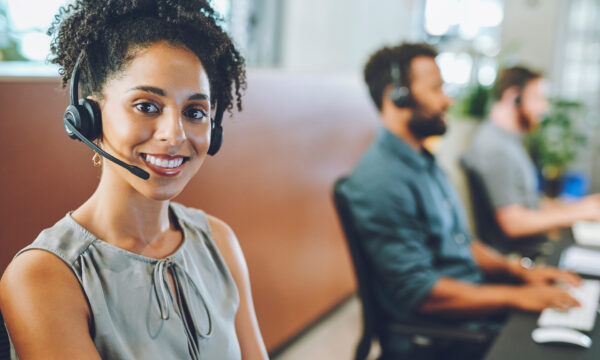 African American female sales team leader or customer service agent working in a call center, talking to a client with a headset. Happy business woman office with diverse colleagues in background.