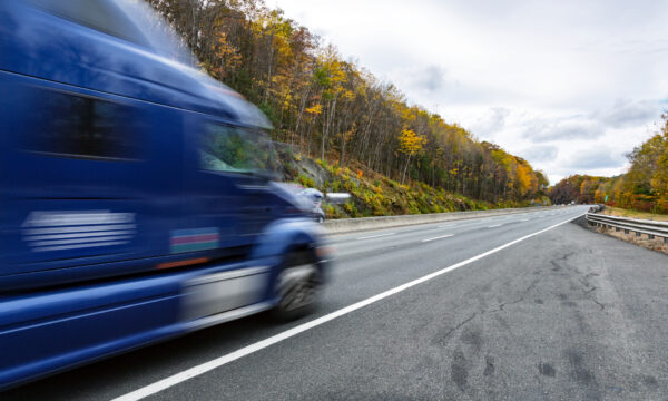A dark blue long exposure motion-blurred semi tractor trailer truck cab rushes past a parked observer under an overcast sky on a bright autumn leaf colored rural USA highway.